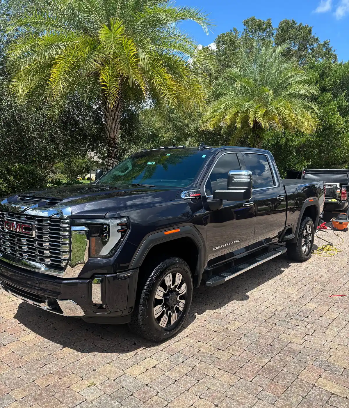Black GMC Sierra HD Denali with a mirror-like finish, parked on a paver driveway under palm trees